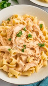 A plate of slow cooker chicken alfredo with fettuccine pasta coated in a rich, creamy parmesan sauce. Shredded chicken is mixed throughout, and the dish is garnished with fresh parsley and extra grated parmesan for added flavor.