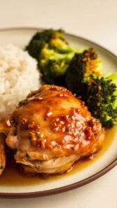 Plated crockpot honey garlic chicken thighs with a glossy honey garlic sauce, served with steamed white rice and roasted broccoli. The chicken is garnished with sesame seeds and chopped green onions, creating a vibrant and appetizing meal on a white ceramic plate.