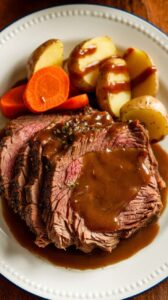 Plated slow cooker pot roast with tender beef, baby potatoes, and whole carrots, drizzled with a rich brown gravy. The hearty meal is served on a white ceramic plate, with warm lighting enhancing the rich colors and textures for an inviting presentation.