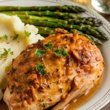 Plated slow cooker garlic butter chicken drizzled with a rich, buttery garlic sauce, served with mashed potatoes and roasted asparagus on a white plate. The golden brown chicken is garnished with fresh parsley, creating an elegant and appetizing meal presentation.