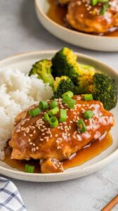 Slow cooker honey garlic chicken served with steamed white rice and roasted broccoli. The chicken is glazed in a thick honey garlic sauce, garnished with sesame seeds and green onions, creating a sweet and savory dish.